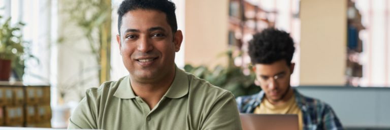 Adult learner smiling in a classroom; another student works on a laptop in the background.