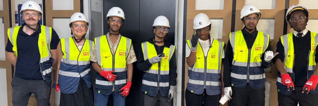 Five ELATT students and two Structure Tone UK staff members pose for a group photo, all wearing hard hats, high-visibility vests, safety glasses, and gloves inside a construction site.