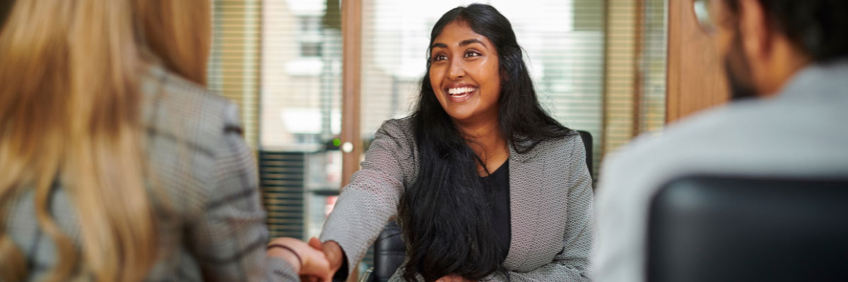 A confident young woman smiling during a job interview, shaking hands with an interviewer in an office setting.