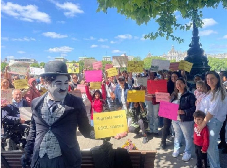 Crowd of people in park facing camera holding signs, foremost sign reads "Migrants need ESOL classes".
