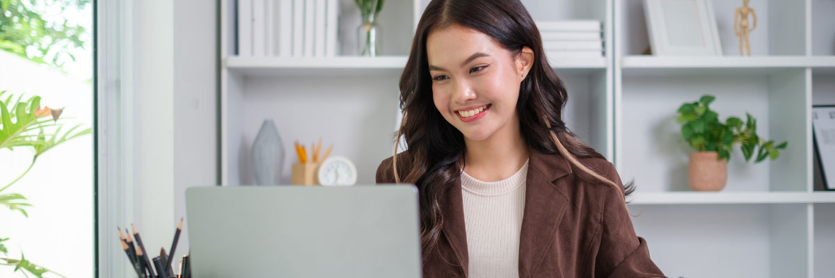 A young woman smiling while working on a laptop in a bright office environment.