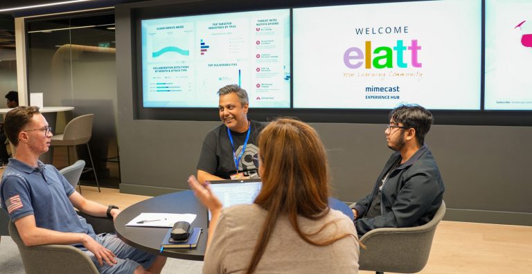 Four people sitting around a table and talking, with large screens on wall behind, largest visible text on screen stating: "WELCOME, elatt, mimecast experience hub".